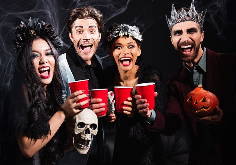 excited multiethnic people with plastic cups laughing at camera during halloween party on black