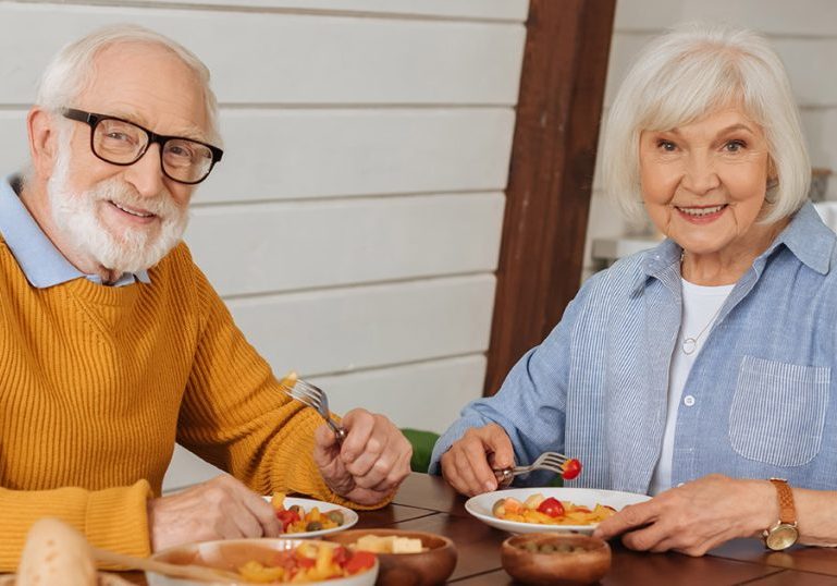 smiling elderly couple looking at camera while sitting at table with vegetarian dinner on blurred foreground in kitchen