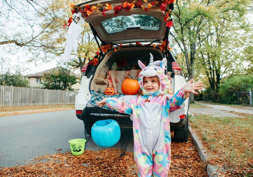 Trick or trunk. Happy baby in unicorn costume celebrating Halloween in trunk of car. Cute smiling toddler preparing for October holiday outdoor. Social distance and safe alternative celebration.